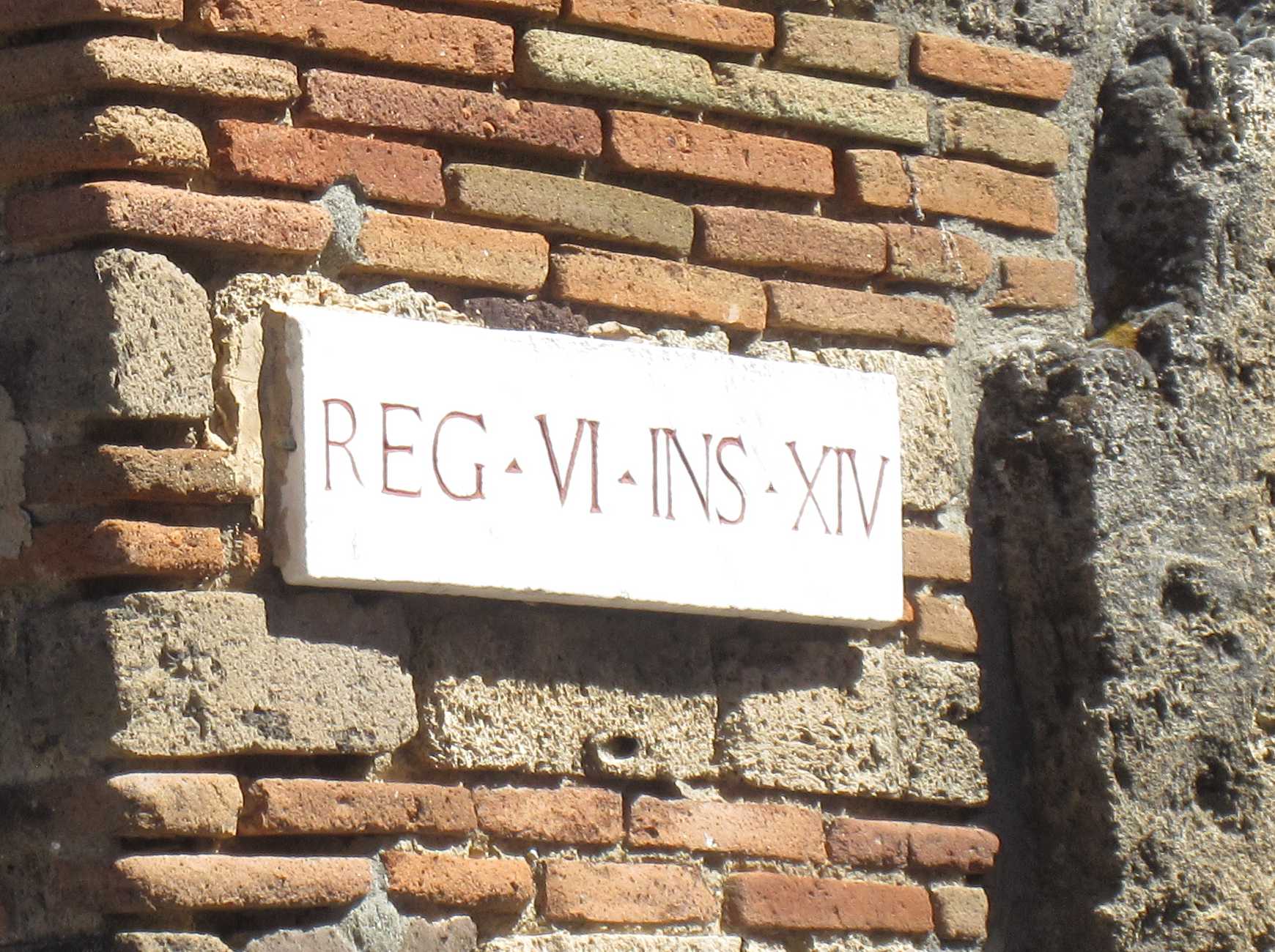 A street sign in Pompeii, saying REG.VI.INS.XIV in red letters engraved and painted on a white marble tile, cemented on a red brick wall.